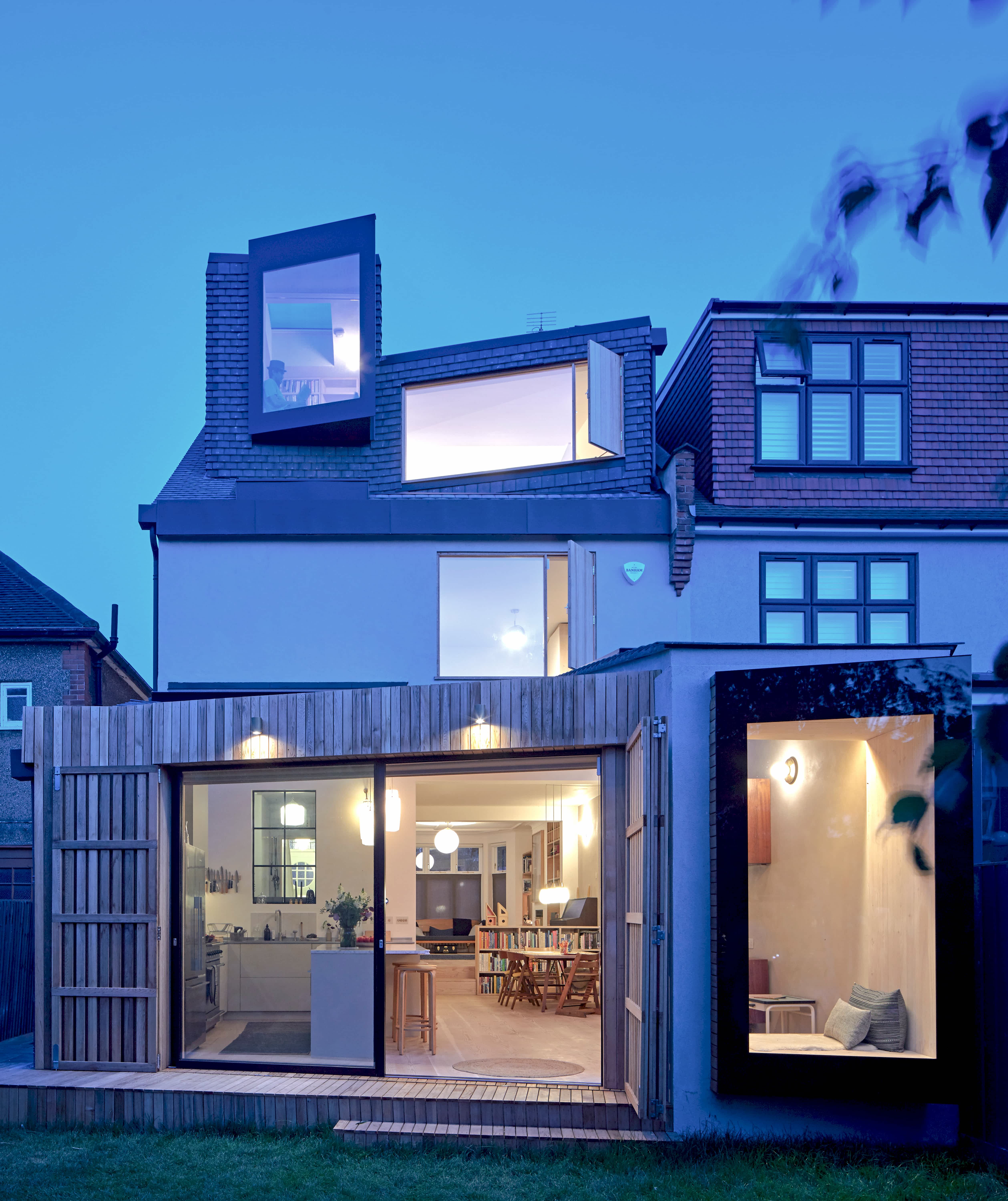 Rear view of Douglas House at dusk, showing open-plan kitchen and dining area with large sliding glass doors, a timber-clad extension, and folding sliding timber shutters designed to reduce solar gain and help keep the home naturally cooler in summer.