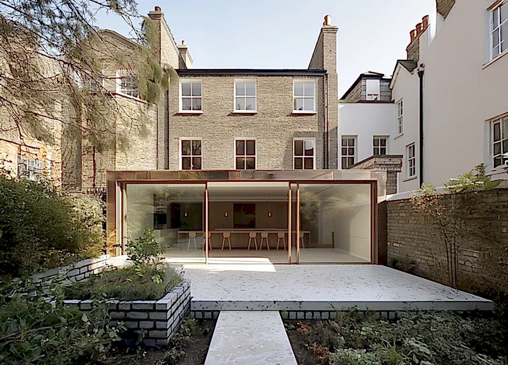 Rear view of a Georgian house with a contemporary copper-clad extension featuring large sliding glass doors, opening onto a minimalist garden terrace designed for low-energy living.