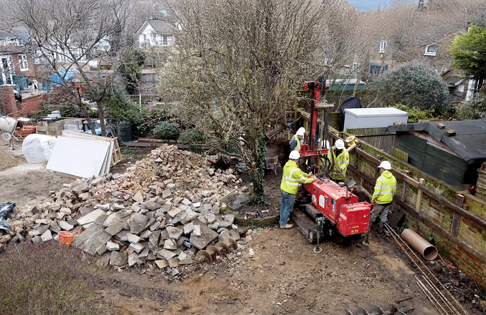 Builders operating piling rig on residential construction site during groundworks, with rubble and foundation materials visible around the area.
