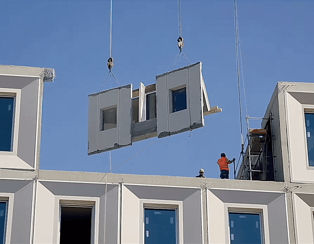 Construction workers guide a prefabricated wall panel with integrated windows into place using a crane, demonstrating Modern Methods of Construction on a multi-storey building.